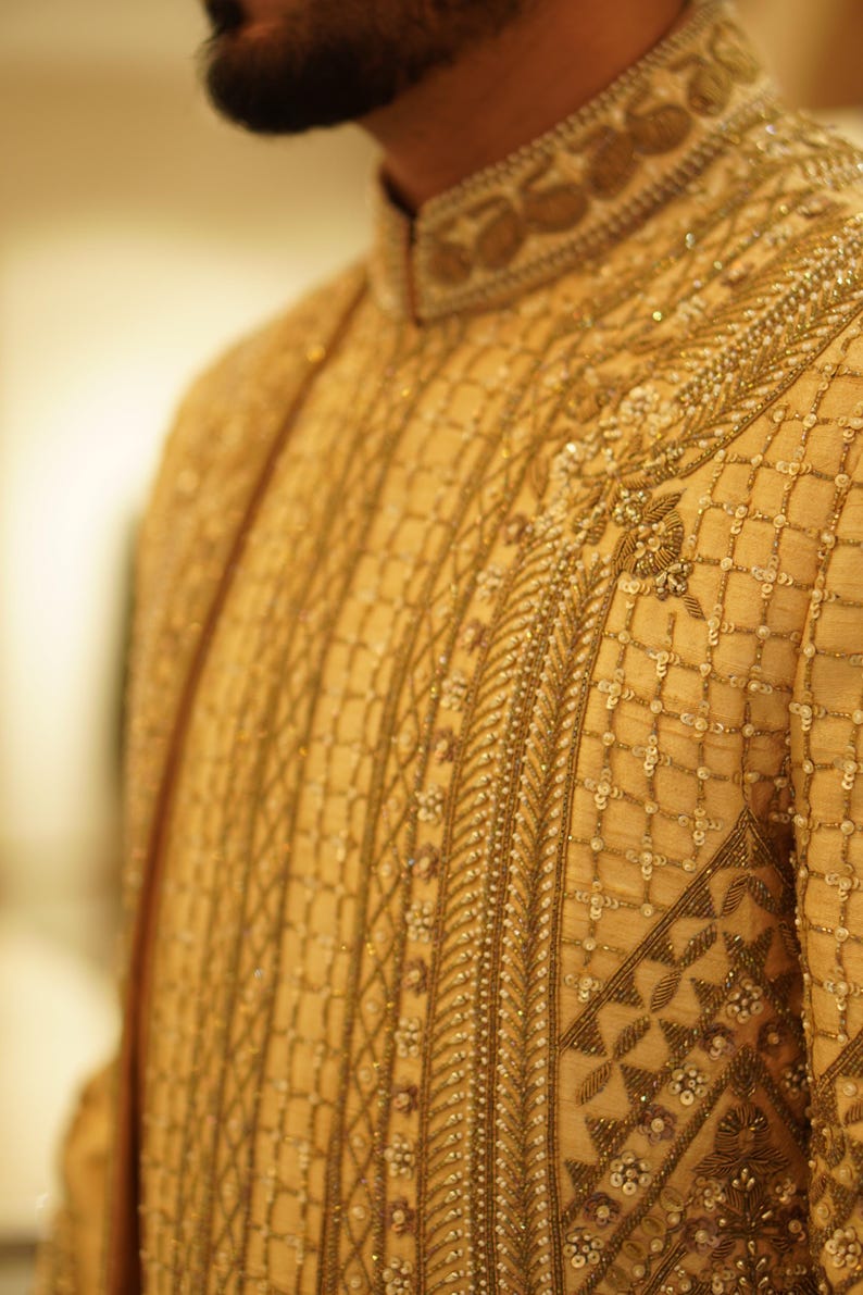K&ouml;nnte beinhalten: Nahaufnahme eines goldenen bestickten Kleidungsst&uuml;cks mit komplizierten Mustern und Perlen. Der Stoff ist hellbeige und die Stickerei ist mit einem dunkleren goldenen Faden ausgef&uuml;hrt.