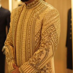 K&ouml;nnte beinhalten: Eine lange, cremefarbene Jacke mit aufwendigen goldenen Stickereien. Die Jacke hat einen hohen Kragen und ein langes, flie&szlig;endes Design. Die Stickereien zeigen geometrische Muster und florale Motive.