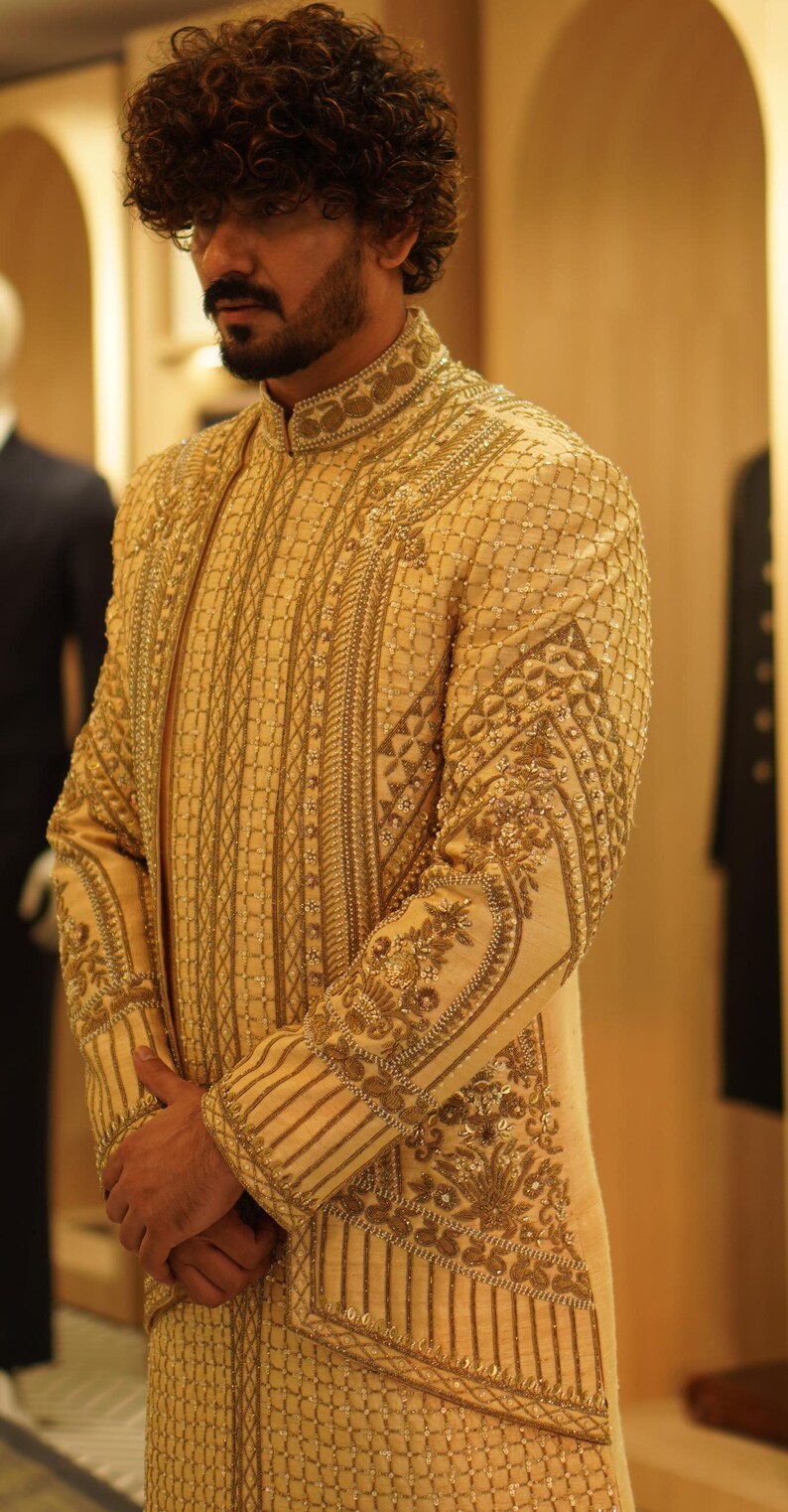 K&ouml;nnte beinhalten: Eine lange, cremefarbene Jacke mit aufwendigen goldenen Stickereien. Die Jacke hat einen hohen Kragen und ein langes, flie&szlig;endes Design. Die Stickereien zeigen geometrische Muster und florale Motive.