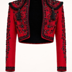 Könnte beinhalten: Eine kurze rote Jacke mit schwarzen Verzierungen und schwarzem Innenfutter. Die Jacke hat aufwendige Perlenarbeiten und dekorative Schulterakzente mit Quasten. Die Ärmel und der Kragen sind ebenfalls mit schwarzen Details versehen.