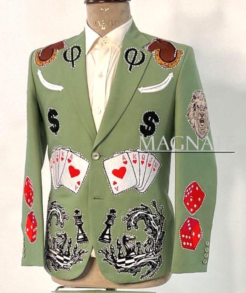 May include: A light green blazer with various embroidered designs, including playing cards, dice, chess pieces, and dollar signs. The blazer also features a lion's head and the word "MAGNATE".