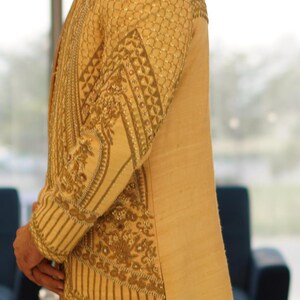 K&ouml;nnte beinhalten: Eine beige, bestickte lange Jacke mit Stehkragen und aufwendigen goldenen Stickereien. Die Jacke wird von einer Person mit lockigem braunen Haar getragen.