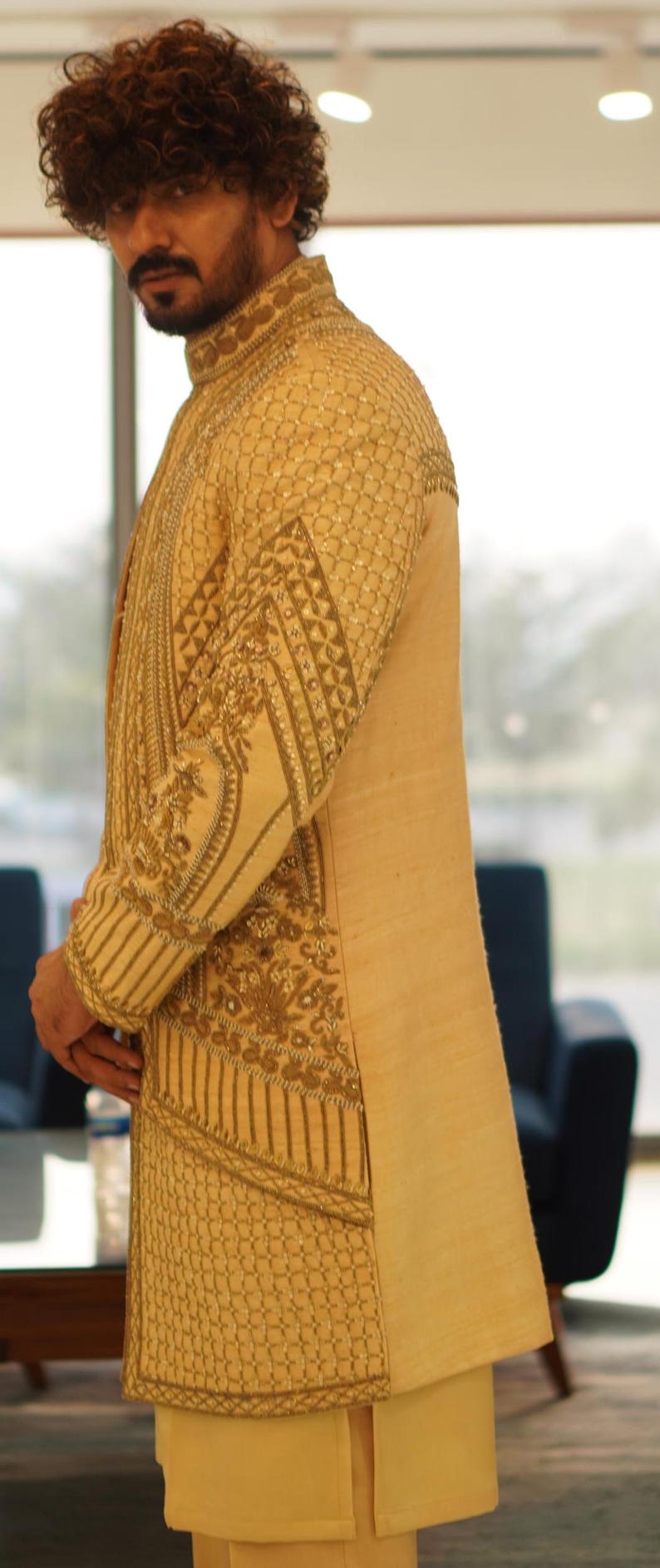 K&ouml;nnte beinhalten: Eine beige, bestickte lange Jacke mit Stehkragen und aufwendigen goldenen Stickereien. Die Jacke wird von einer Person mit lockigem braunen Haar getragen.