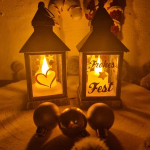 Könnte beinhalten: Zwei dekorative Laternen mit brennenden Kerzen, eine mit Herz und Sternen, die andere mit dem Text "Frohes Fest". Goldene Weihnachtskugeln befinden sich vor den Laternen. Der Hintergrund umfasst Zuckerstangen und eine Weihnachtsmann-Dekoration.