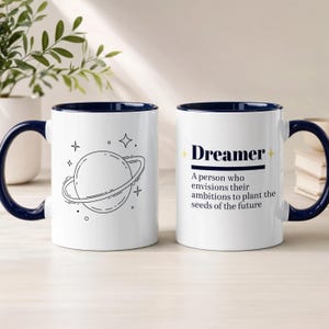 May include: Two white ceramic mugs with dark blue interiors and handles. One mug features a black line art illustration of a planet with rings and stars. The other mug displays the text Dreamer and the definition: A person who envisions their ambitions to plant the seeds of the future.