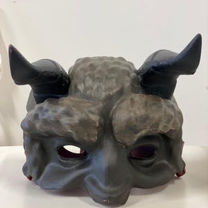 Può includere: Una maschera in cartapesta nera e marrone con corna e una superficie testurizzata. La maschera ha due fori per gli occhi e una bocca larga.