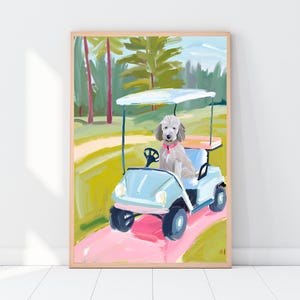 Op de afbeelding: Een ingelijste kunstdruk van een hond in een golfkar. De hond is lichtgrijs met een roze halsband. De golfkar is lichtblauw met een wit dak, op een roze pad. De achtergrond toont groene bomen en gras.