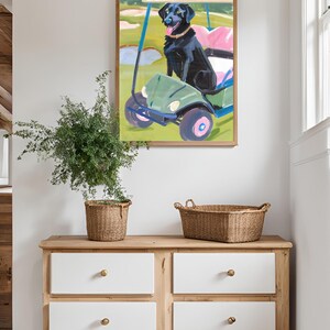 Puede incluir: Una pintura enmarcada de un perro negro sentado en un carrito de golf verde en un campo de golf. El perro lleva un collar rosa y tiene la lengua fuera.