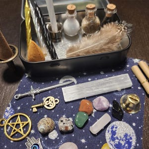 May include: A collection of spiritual items, including candles, small bottles, feathers, crystals, and charms, arranged on a dark blue cloth with a celestial pattern. The items are in a small metal tin.