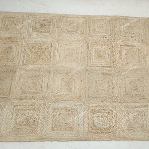 Może przedstawiać: Prostokątny dywan z juty z geometrycznym wzorem kwadratów. Dywan jest w naturalnym beżowym kolorze i wydaje się być tkany ręcznie. Kwadraty są równomiernie rozmieszczone na powierzchni dywanu, tworząc wzór przypominający siatkę. Odpowiedni do dekoracji wnętrz.