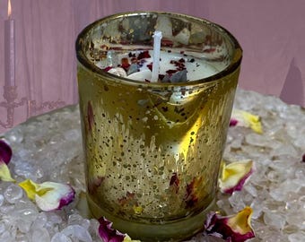 May include: A gold glass candle holder with a white candle and dried flowers and crystals inside. The candle holder is sitting on a bed of white crystals.