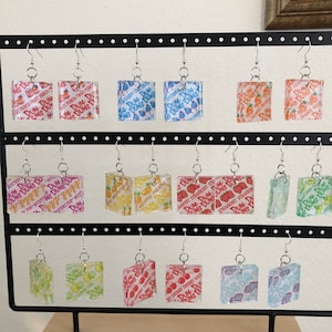 May include: A set of 16 pairs of earrings featuring square-shaped charms with various Dum Dums lollipop flavors. The charms are made of clear plastic with colorful graphics and text. The earrings are displayed on a black stand.