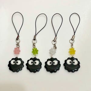 以下が含まれることがあります： 異なる色のビーズが付いた、黒と白のキャラクターチャームが4つ。チャームは黒で、白い目が付いており、波打つような輪郭をしています。ビーズはピンク、グリーン、ホワイト、イエローです。