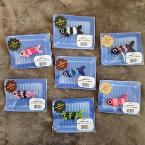 Peut inclure: Sept ornements de poisson au crochet de différentes couleurs, chacun emballé sur un petit plateau en plastique bleu avec une étiquette faite main. Les poissons présentent des motifs rayés et des yeux noirs, avec des étiquettes 100% FRESH FISH.