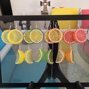 Op de afbeelding: Een collectie oorbellen met fruitmotieven, waaronder citroen, limoen, sinaasappel en grapefruit schijfjes, tentoongesteld op een zwart gaasrek. De oorbellen zijn gemaakt van een doorschijnend materiaal. Elke paar hangt aan een zilveren haak.