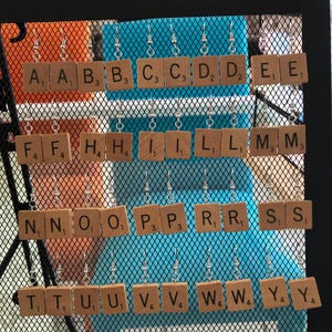 May include: A set of wooden letter tile earrings, each tile featuring a different letter of the alphabet. The tiles are displayed on a wire rack with a blue background.