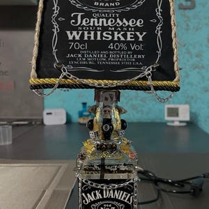 Può includere: Una lampada personalizzata realizzata con una bottiglia di whisky Jack Daniel's. La lampada presenta un paralume nero con finiture dorate e il logo Jack Daniel's. La bottiglia è decorata con dettagli in metallo e una catena. Contiene 70cl.