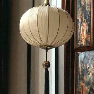 May include: A white, pleated, round lampshade hangs from a gold cord with a black tassel. The lampshade is suspended from a gold ceiling fixture. The lamp is hanging in front of a wooden framed picture with a floral design.