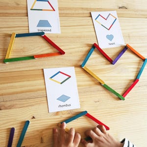 May include: A child's hand builds geometric shapes using colorful craft sticks.  The shapes are matched to cards with the names "trapezoid", "heart", and "rhombus".