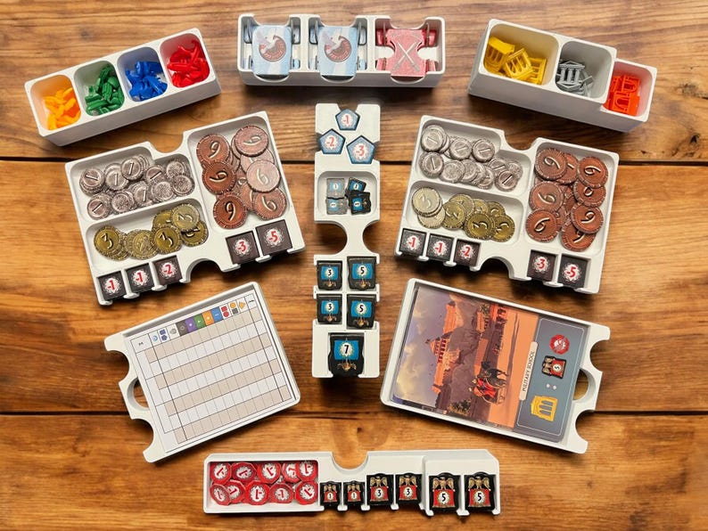 May include: A white plastic organizer with compartments for storing game pieces. The organizer has a variety of compartments, including some with dividers. The compartments contain a variety of game pieces, including coins, tokens, and cards. The organizer is designed to help players keep their game pieces organized and easy to access.