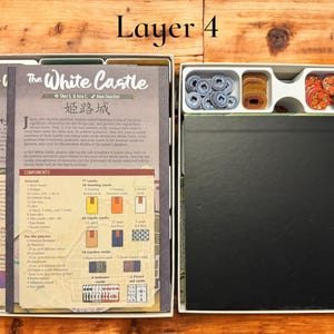May include: The White Castle board game box, layer 4, showing the game board, rule book, and components in a white box with a black lid. The components include cards, tokens, and dice.