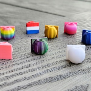Op de afbeelding: Een verzameling kleine, kleurrijke, 3D-geprinte decoratieve objecten. De objecten omvatten bollen en geometrische vormen in verschillende kleuren, waaronder roze, geel, blauw en een regenboogverloop. Ze zijn gerangschikt op een houten oppervlak.