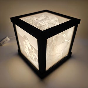 May include: A black cube-shaped lamp with a white light shining through a translucent image of two people. The lamp is on a white surface.