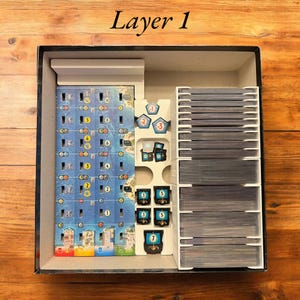May include: A white organizer insert for a board game box. The insert has compartments for storing game pieces, cards, and a game board. The insert is labeled "Layer 1".