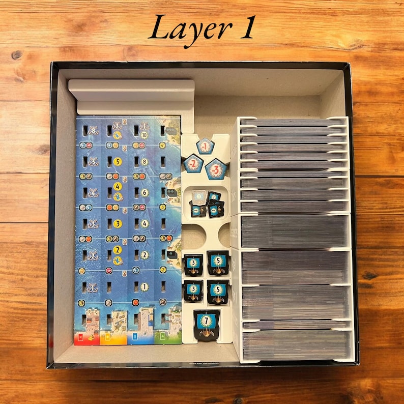May include: A white organizer insert for a board game box. The insert has compartments for storing game pieces, cards, and a game board. The insert is labeled "Layer 1".