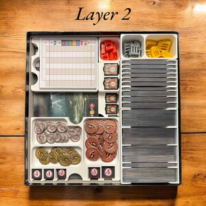 May include: A white plastic organizer with compartments for storing game pieces. The organizer has a label that says "Layer 2". The compartments contain small plastic tokens, dice, and cards.