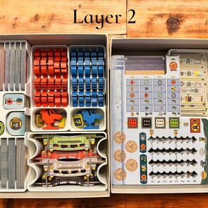 May include: A white plastic organizer with multiple compartments for storing game pieces. The organizer is labeled "Layer 2". The compartments contain various game pieces, including small wooden cubes, plastic tiles, and cards.