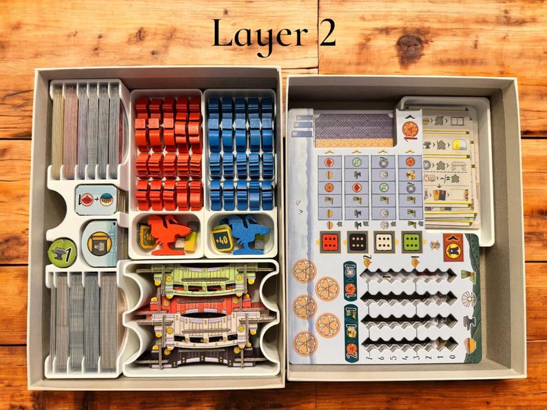 May include: A white plastic organizer with multiple compartments for storing game pieces. The organizer is labeled "Layer 2". The compartments contain various game pieces, including small wooden cubes, plastic tiles, and cards.