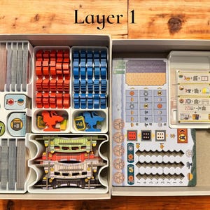 May include: A white plastic organizer with multiple compartments for storing game pieces. The organizer is labeled "Layer 1". The compartments contain various colored plastic pieces, dice, and cards.
