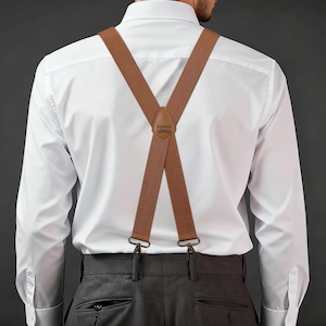 Puede incluir: Tirantes marrones con cierres metálicos, usados sobre una camisa blanca y pantalones grises. Los tirantes tienen un parche de cuero con la palabra "Spoon" y un diseño de bigote. Un accesorio clásico.