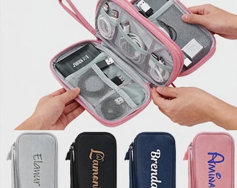 Può includere: Organizzatore da viaggio rosa con cerniera e più scomparti, contenente vari accessori elettronici come cavi e un power bank. Sotto, quattro custodie più piccole in grigio, nero, blu navy e rosa, ciascuna con un nome ricamato.