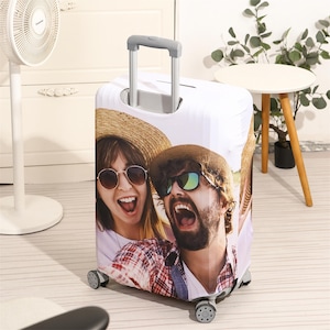 May include: A white suitcase cover with a colourful image of two people wearing sunglasses and straw hats, smiling. The luggage has a retractable handle and four wheels, ideal for travel.