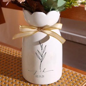 Peut inclure: Un vase en céramique blanc avec un bord festonné, noué avec un ruban doré. Le vase présente une illustration botanique noire et les initiales "j + h" avec la date "02.15.25" imprimée en dessous. Le vase est rempli de fleurs.