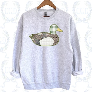 May include: A pale grey sweatshirt with a decorative duck design. The duck is made from various patterned fabrics in shades of green, brown, and white, with a yellow beak and a green plaid head. The sweatshirt has a crew neck and long sleeves.