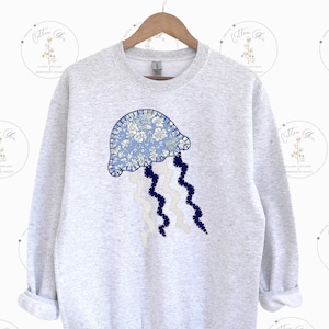 Könnte beinhalten: Ein grauer Pullover mit einem blauen und weißen Blumen-Quallen-Applikation. Die Qualle hat einen blauen Körper mit weißen Blumen-Details und weiße Tentakel mit blauem Besatz.