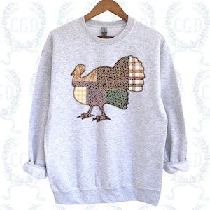 Könnte beinhalten: Ein hellgraues Sweatshirt mit einem Patchwork-Truthahn-Design. Der Truthahn besteht aus verschiedenen gemusterten Stoffen in Braun-, Gelb- und Grüntönen. Das Sweatshirt wird auf einem Holzbügel präsentiert.