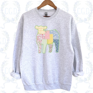 May include: A light gray sweatshirt featuring a patchwork sheep design. The sheep is composed of various fabric patterns, including floral, gingham, and patterned blue. The sweatshirt has a crew neck and long sleeves, and is hanging on a wooden hanger.