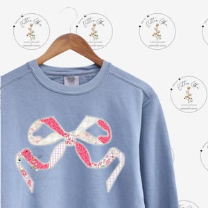 May include: A light blue sweatshirt with a large pink and white patchwork bow applique. The bow is made of different fabrics with floral and geometric patterns.