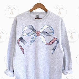 Puede incluir: Una sudadera gris con un gran lazo de retazos bordado en la parte delantera. El lazo está hecho de diferentes telas en tonos azules, rojos y blancos.