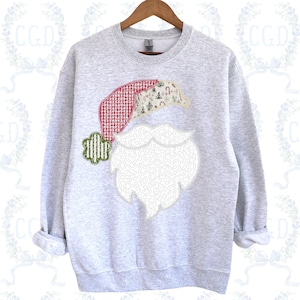 May include: A light gray sweatshirt featuring a Santa Claus design. The design includes a red and white checkered hat, a white beard, and a green and white striped embellishment. The hat has a festive pattern with trees and candy canes.