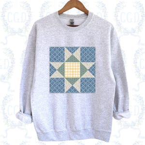 May include: A light gray sweatshirt featuring a colorful quilt block design. The design includes blue, green, and cream-colored star shapes and a yellow gingham center. The sweatshirt has a crew neck and long sleeves.