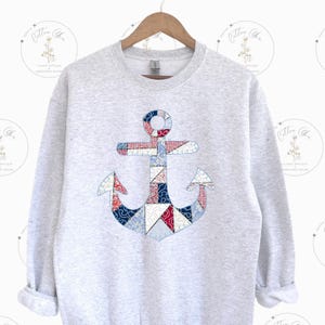 Puede incluir: Una sudadera gris claro con un gran aplique de ancla. El ancla está hecha de tela de retazos con patrones rojos, blancos y azules. La sudadera tiene cuello redondo y mangas largas.
