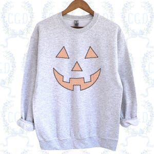 Puede incluir: Una sudadera gris claro de cuello redondo con un gráfico de cara de calabaza. Los ojos, la nariz y la boca dentada de la calabaza están rellenos con un patrón de cuadros vichy naranja y blanco y delineados con costuras oscuras en el frente.