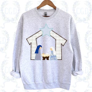 May include: A light gray sweatshirt featuring a nativity scene. The design includes a star, a stable, and figures of Mary and Joseph. The figures are in blue and white patterned fabrics. The sweatshirt is on a wooden hanger.