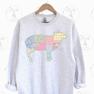 Puede incluir: Una sudadera gris con un diseño de oveja de patchwork. La oveja está hecha de diferentes cuadrados de tela floral y a cuadros.