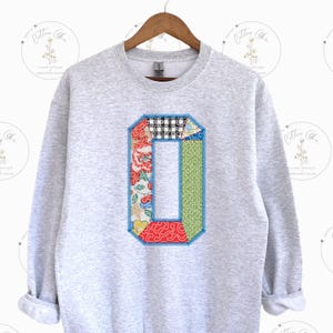 May include: A gray sweatshirt with a large patchwork letter "O" on the front. The letter is made of different fabrics and colors, including red, green, blue, and white.